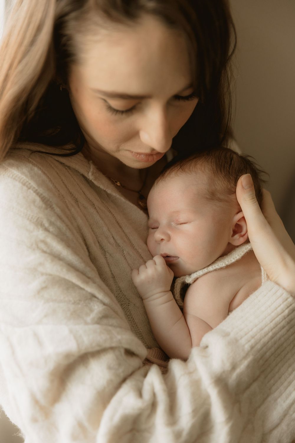 A tender moment of parent and newborn baby cuddling in warm lighting while wearing a cozy white sweater.