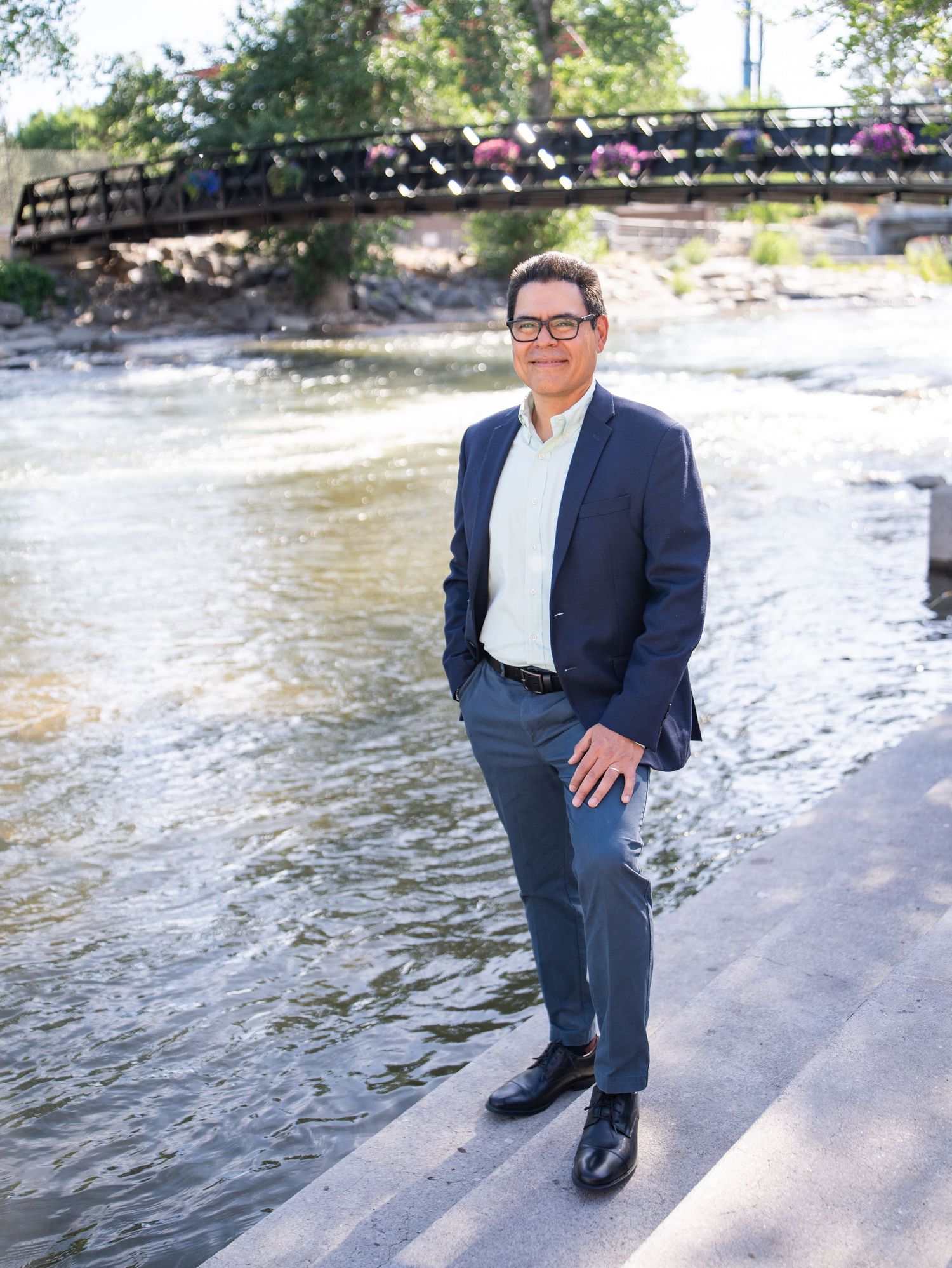 Well-dressed person in a navy suit and glasses stands by a river with a footbridge in the background on a sunny day.