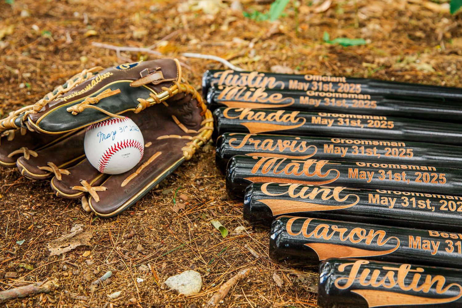 A leather baseball glove with a baseball next to black wooden bats with handwritten names on them laying on dirt ground.