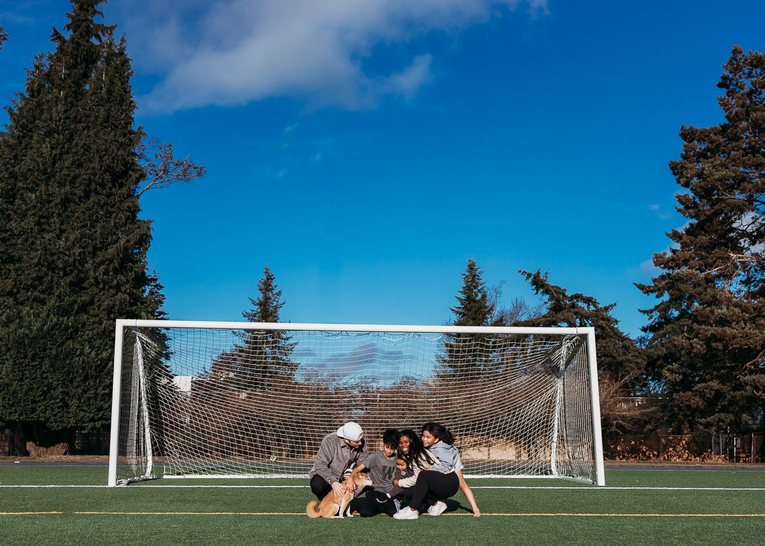 People sitting together on a soccer field in front of a goal post surrounded by tall pine trees against a blue sky.