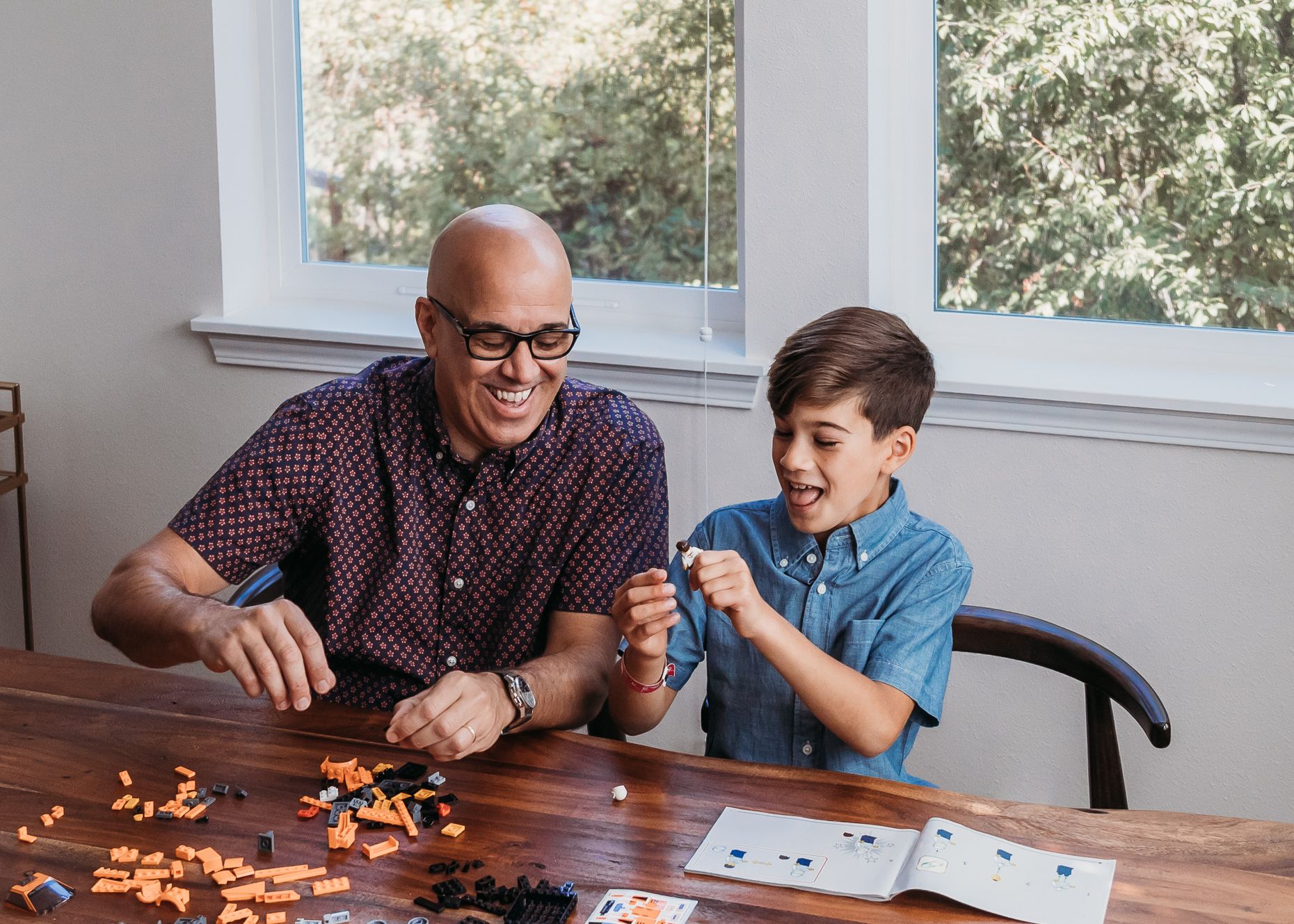 A man and young boy sit at a table working together on an activity with colorful pieces spread out in front of them.