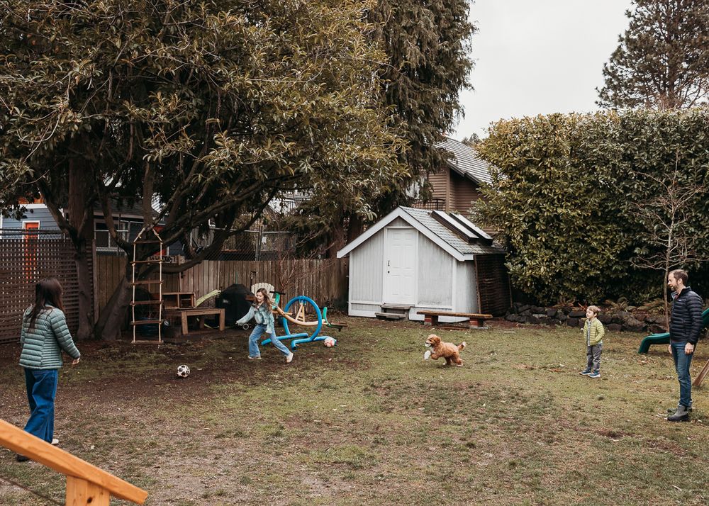 Family playing together in a backyard with swings, trees and a small white shed.