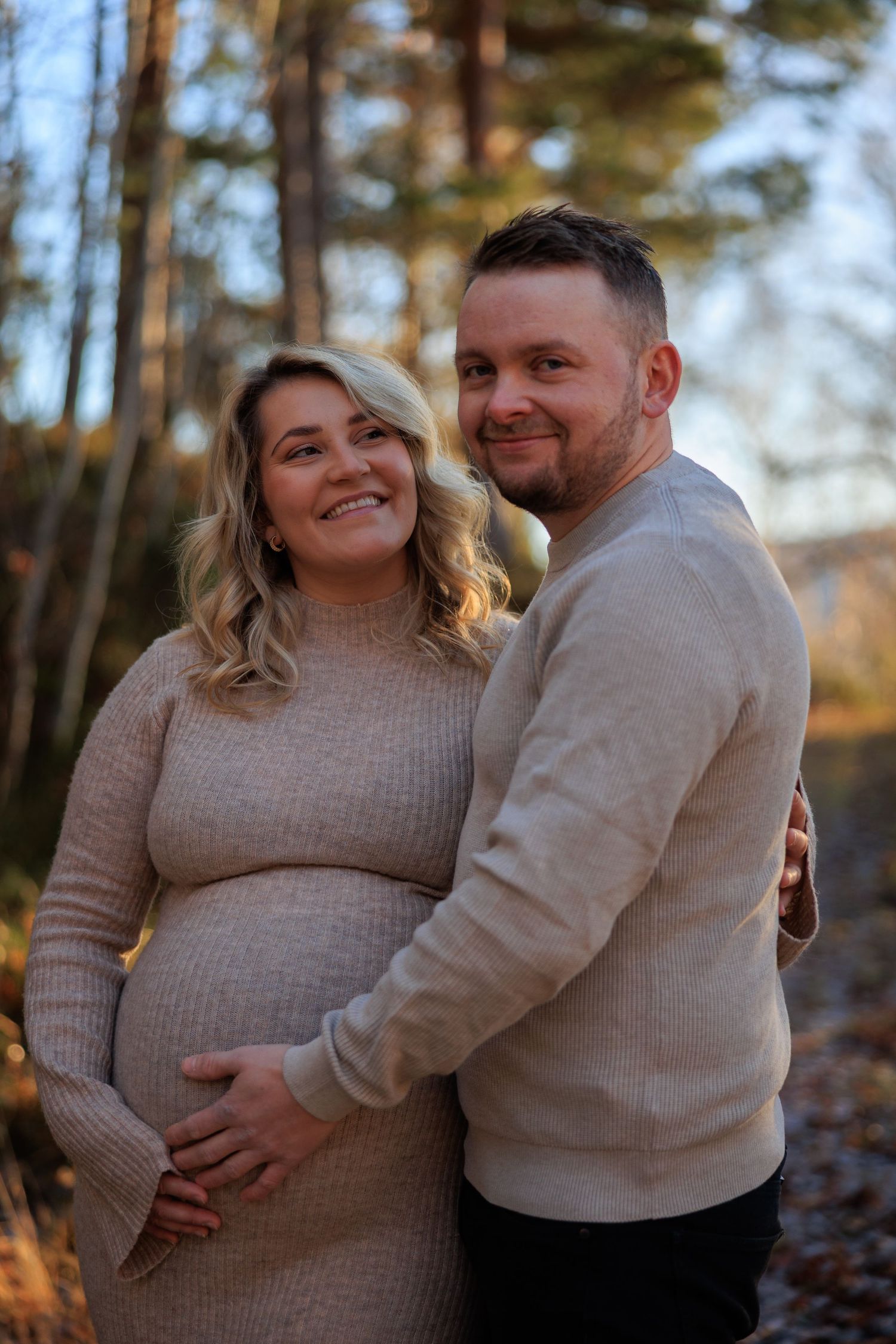 A couple in matching beige sweaters embrace during an outdoor maternity photoshoot in an autumn forest setting.