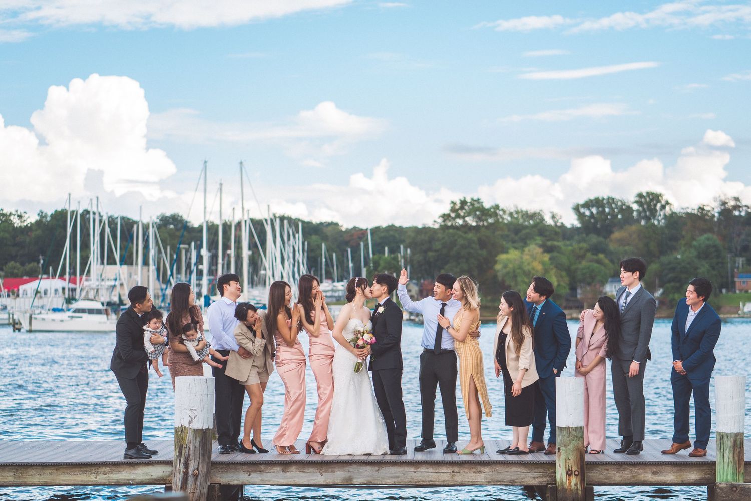 Wedding party and guests gathered on a dock with sailboats in the marina background.
