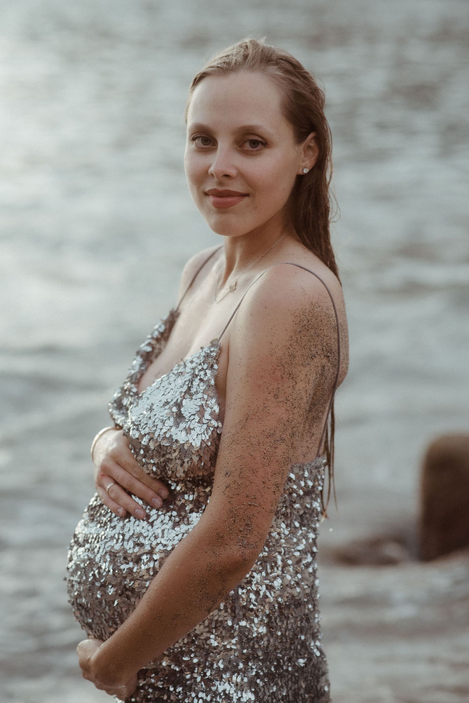 A woman in a sparkly dress poses by the water in a moody, overcast setting.