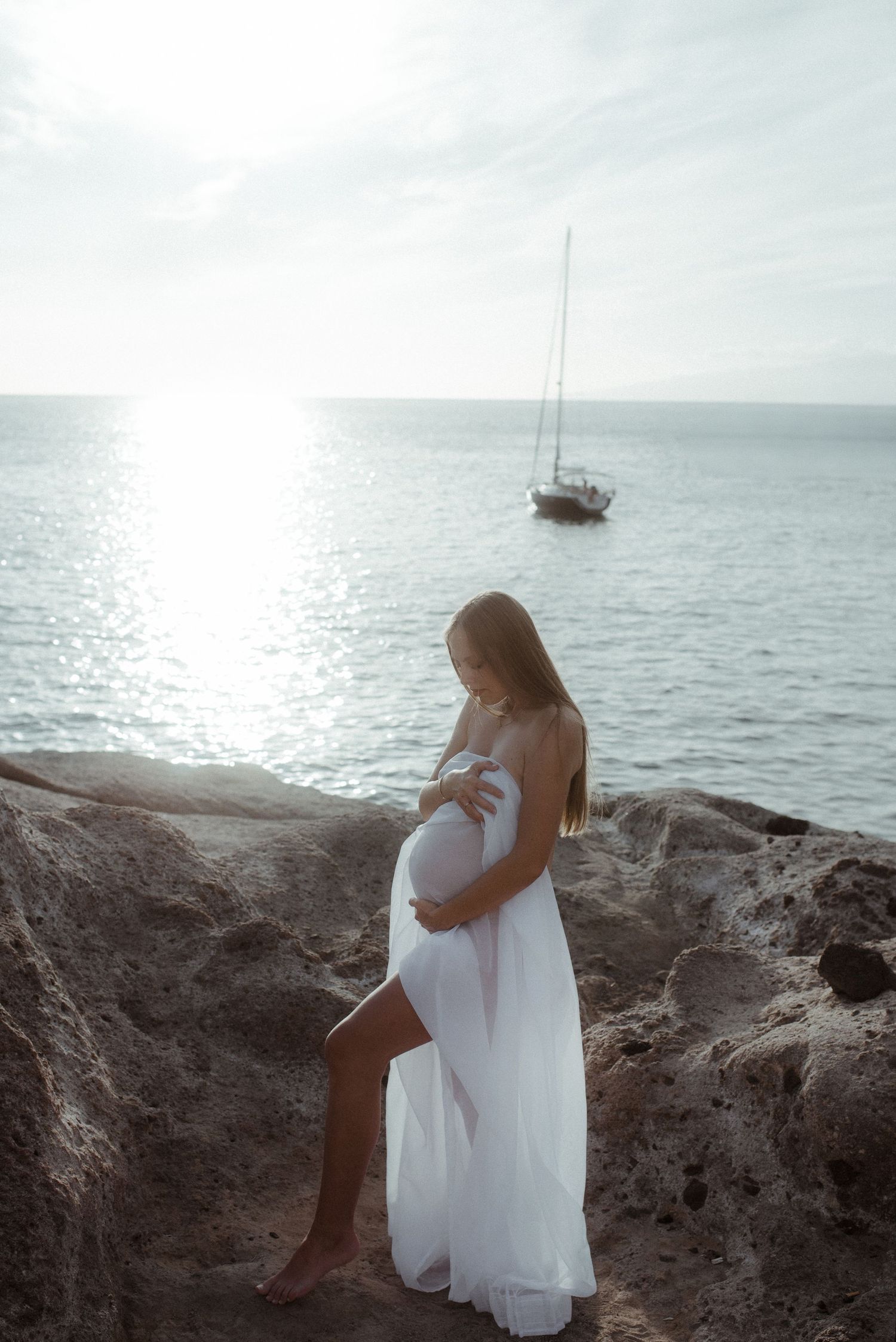 Ethereal maternity photoshoot on rocky coastline at sunset with sailboat in distance.