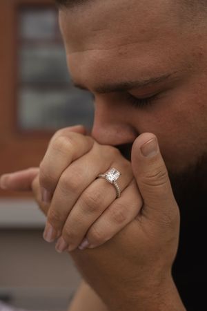 Person with a beard covers their face emotionally while wearing an engagement ring.