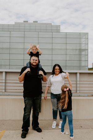 Family members engage in playful interactions outdoors against a building wall.