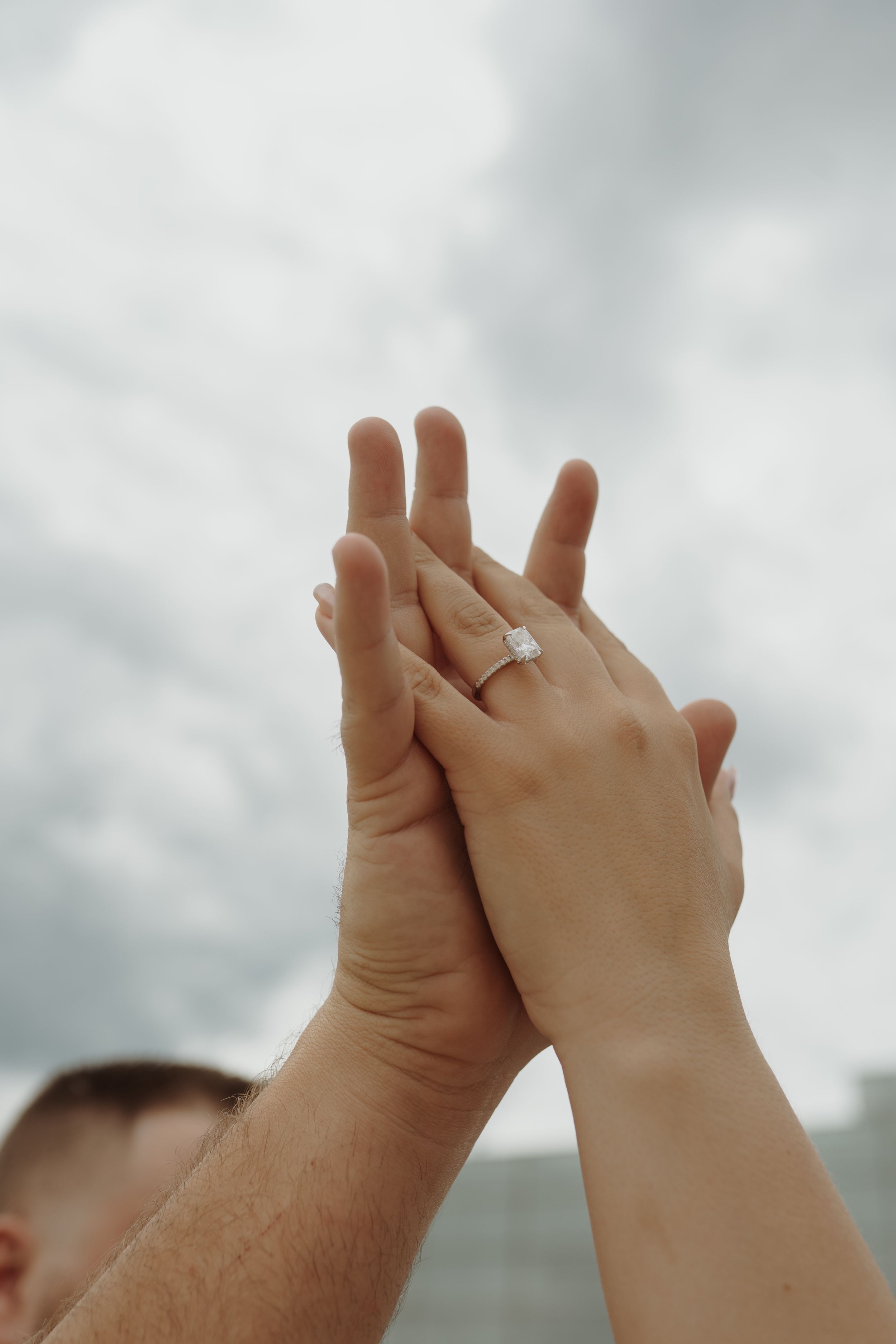 Two hands intertwined against a cloudy sky, with an engagement ring visible.