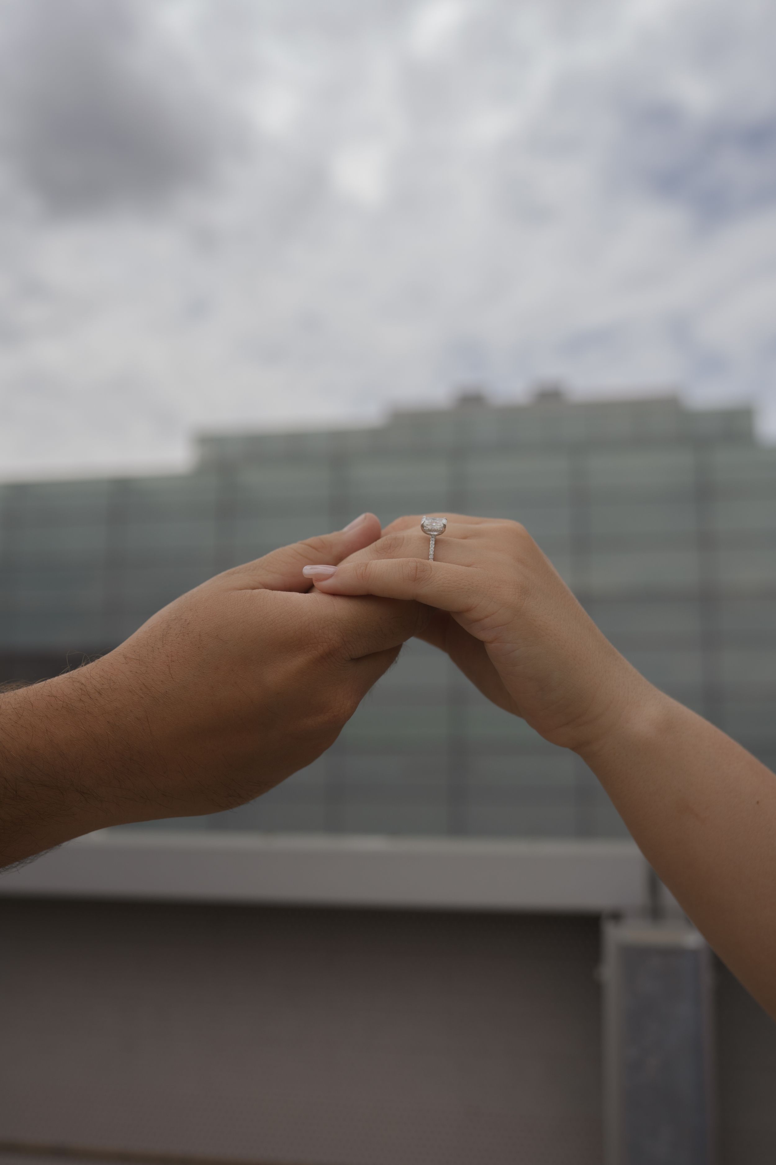 Two hands reaching towards each other with an engagement ring visible against a cloudy backdrop.