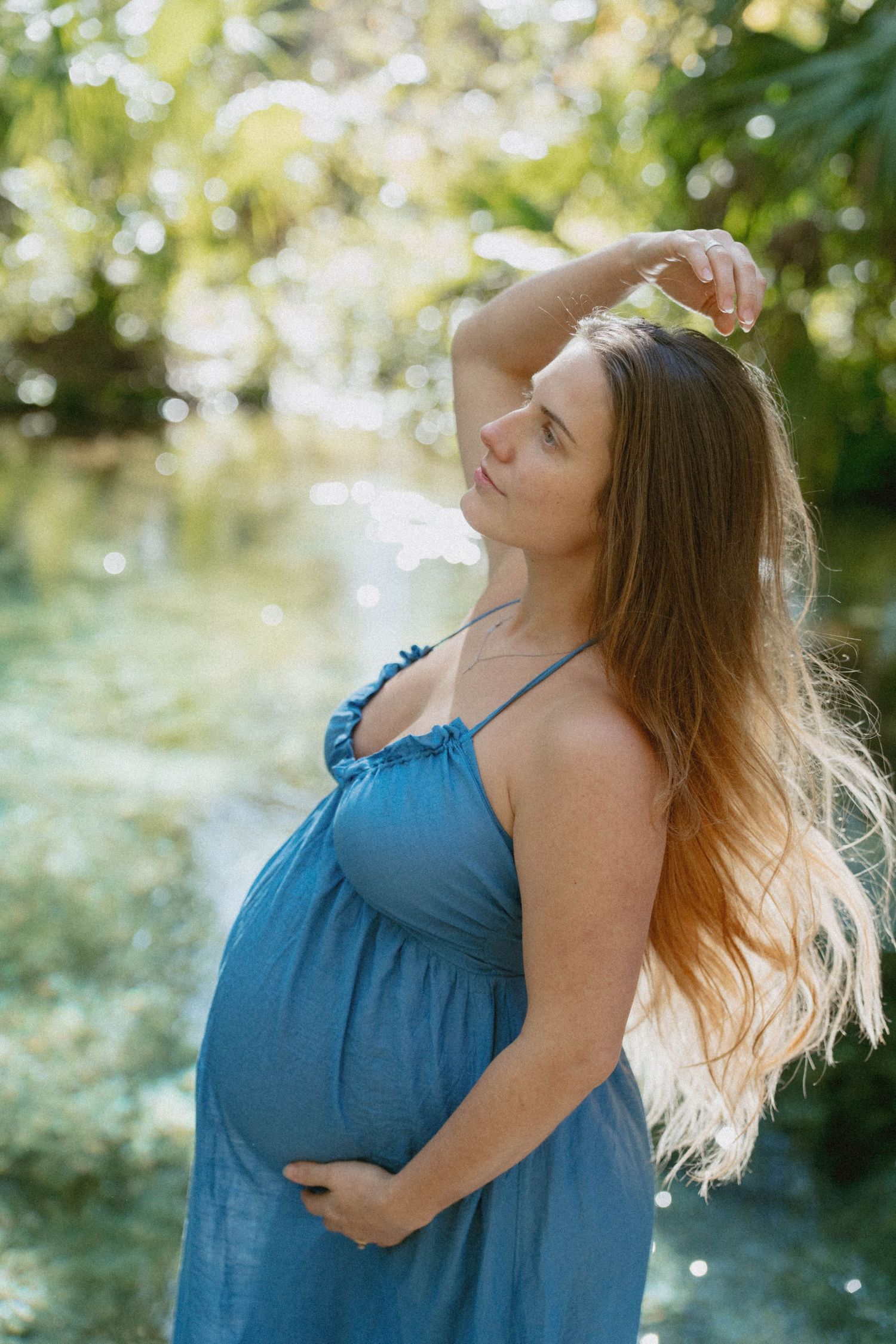 A pregnant woman in a flowing blue sundress stands in dappled sunlight with her hand gracefully raised.