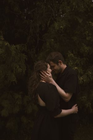 A tender embrace captured in dark, moody lighting against forest foliage.