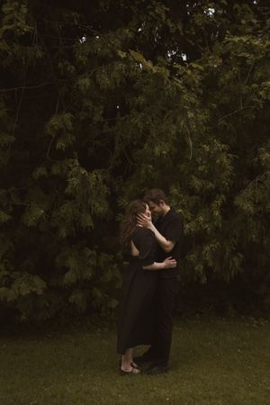 A couple in black clothing sharing a romantic moment on grass near trees.