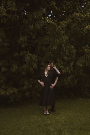 Two people in black attire embracing on grass with trees in background.