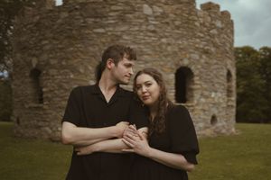 A couple embraces near an ancient stone castle wall on an overcast day.