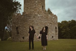Two people in black clothing stand near historic stone ruins on a grassy hill.