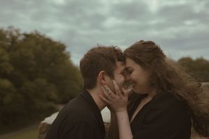 A romantic couple in black outfits share an intimate moment against a moody cloudy sky.