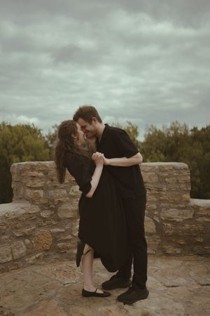 Two people in dark clothing share an emotional moment near a stone wall.