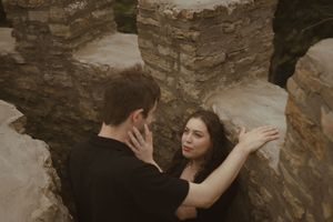 Two people share an intimate moment against stone walls and cloudy skies.