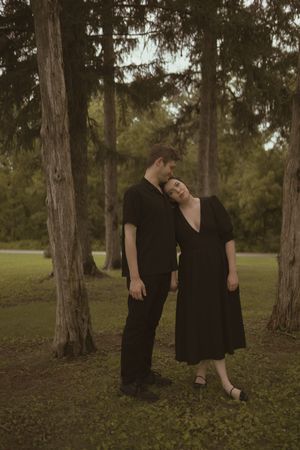 Two people in black clothing dancing together among tall pine trees in a forest setting.
