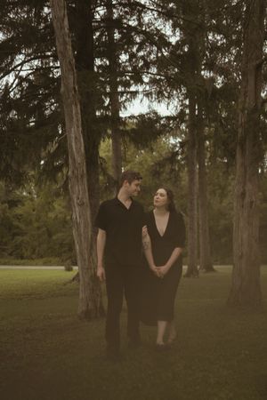 Two people in dark clothing share a tender moment among pine trees at dusk.