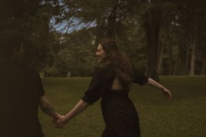 Two people in black clothing dance and laugh together in a wooded park.
