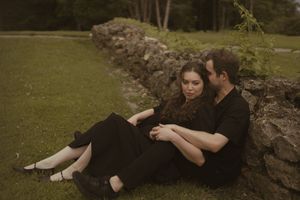Two people dressed in dark clothing embrace while sitting near a stone wall outdoors.