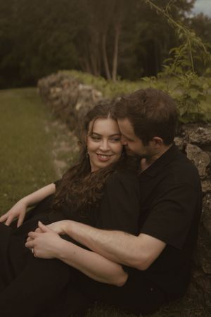 Two people in dark clothing embrace lovingly against a natural outdoor background.