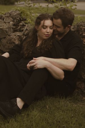 A couple dressed in black shares intimate poses near stone wall in an outdoor setting.