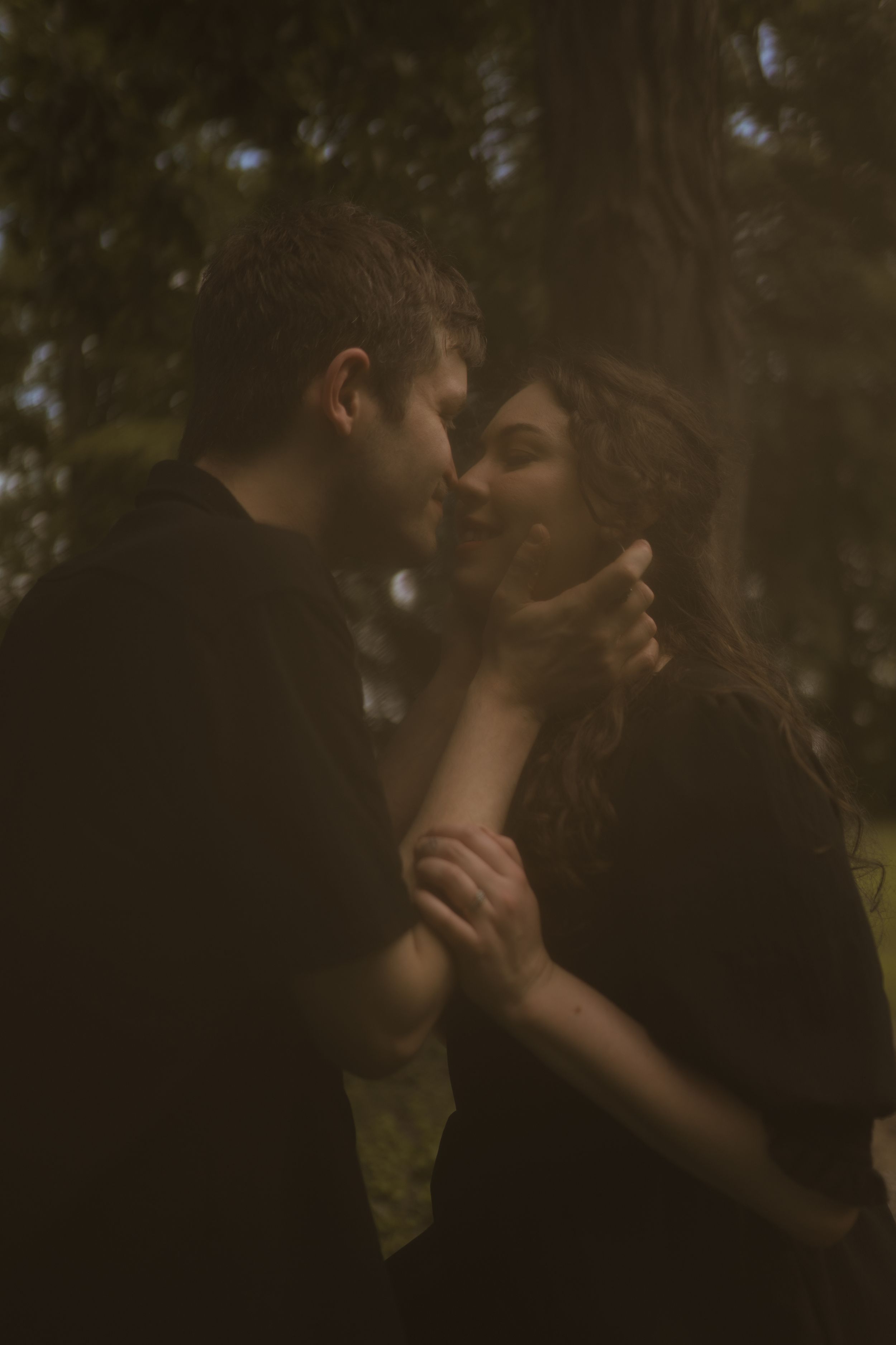 A tender moment captured between two people in a shadowy forest setting.