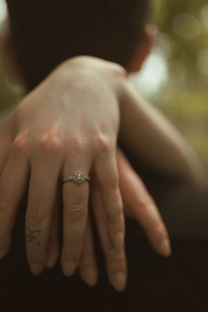 A close-up view of hands showing an engagement ring against a blurred background.