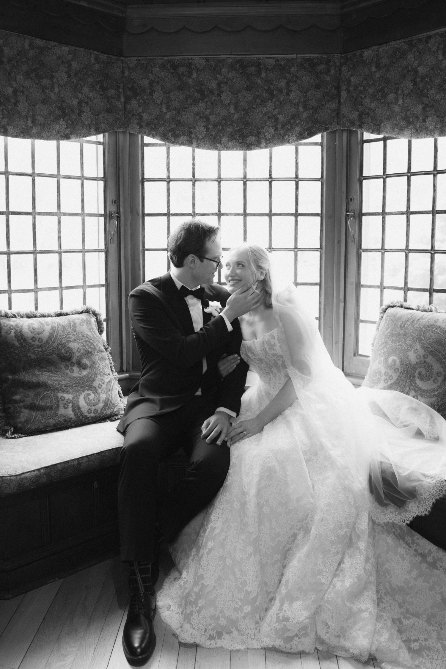 Black and white wedding photo of bride and groom sharing a tender moment by a bay window on a cushioned bench.