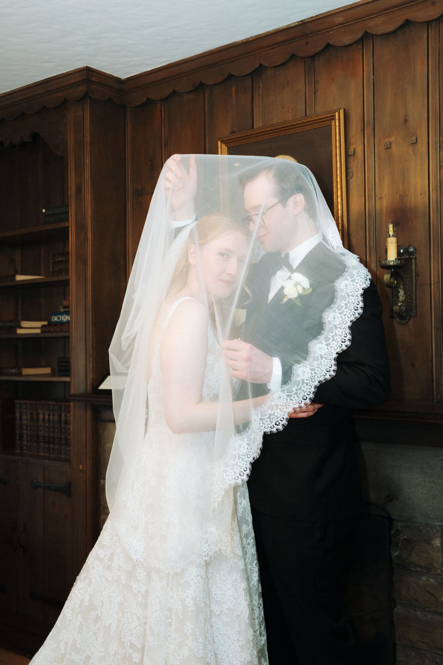 Bride and groom share intimate moment under lace wedding veil in wood-paneled room.