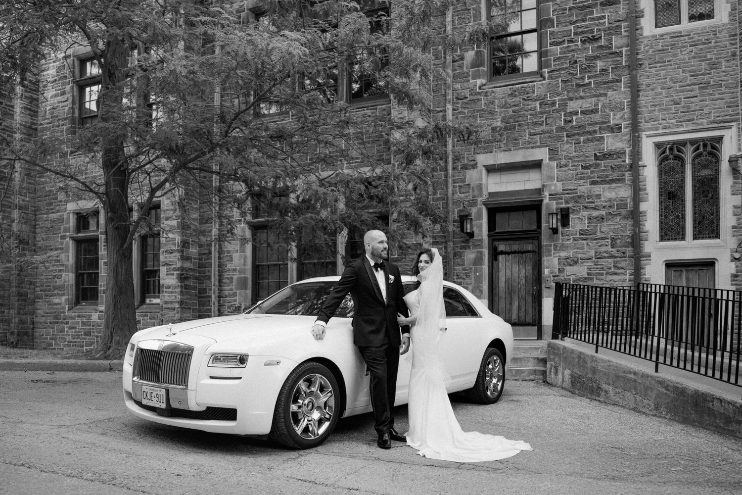 Bride and groom posing beside a white Rolls-Royce Ghost outside a stone building on their wedding day.