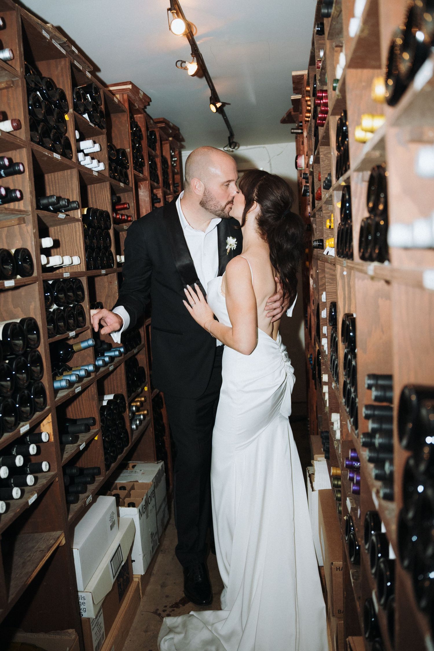 Bride and groom sharing a romantic kiss in a wine cellar, surrounded by wooden wine racks filled with bottles.