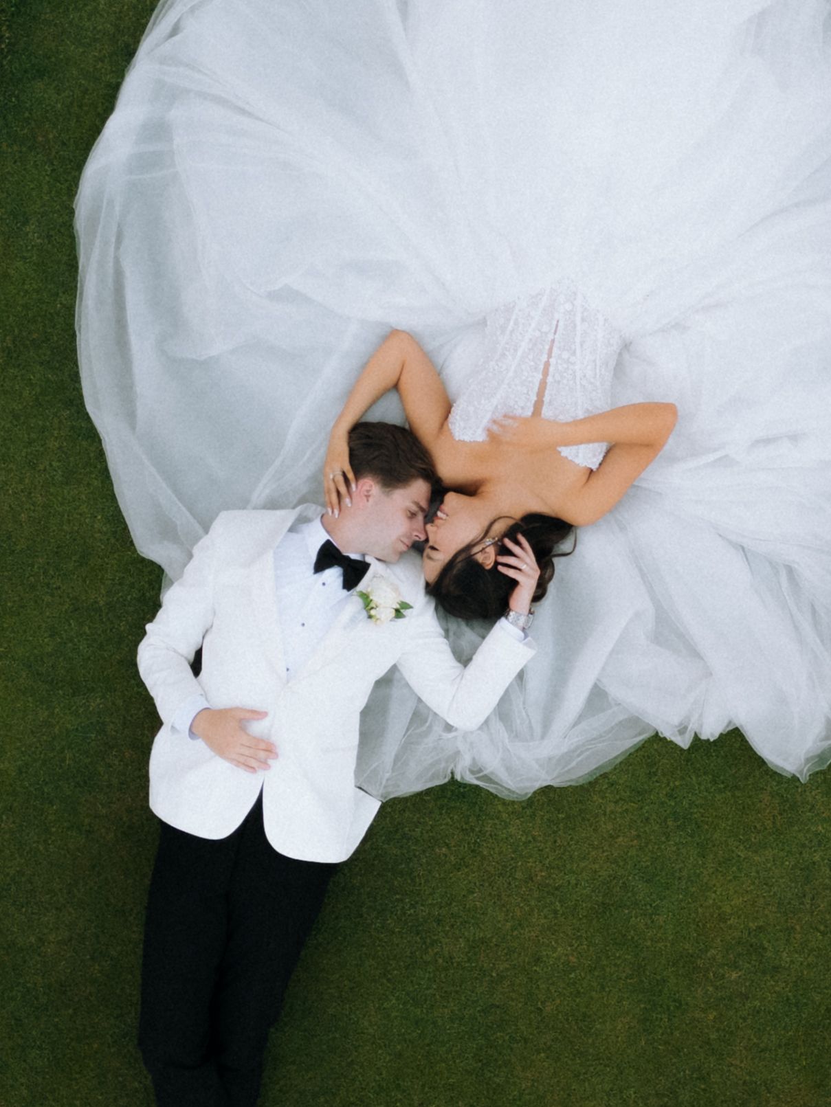 Aerial view of newlyweds lying on green grass, bride in white tulle gown and groom in white tuxedo sharing a kiss.