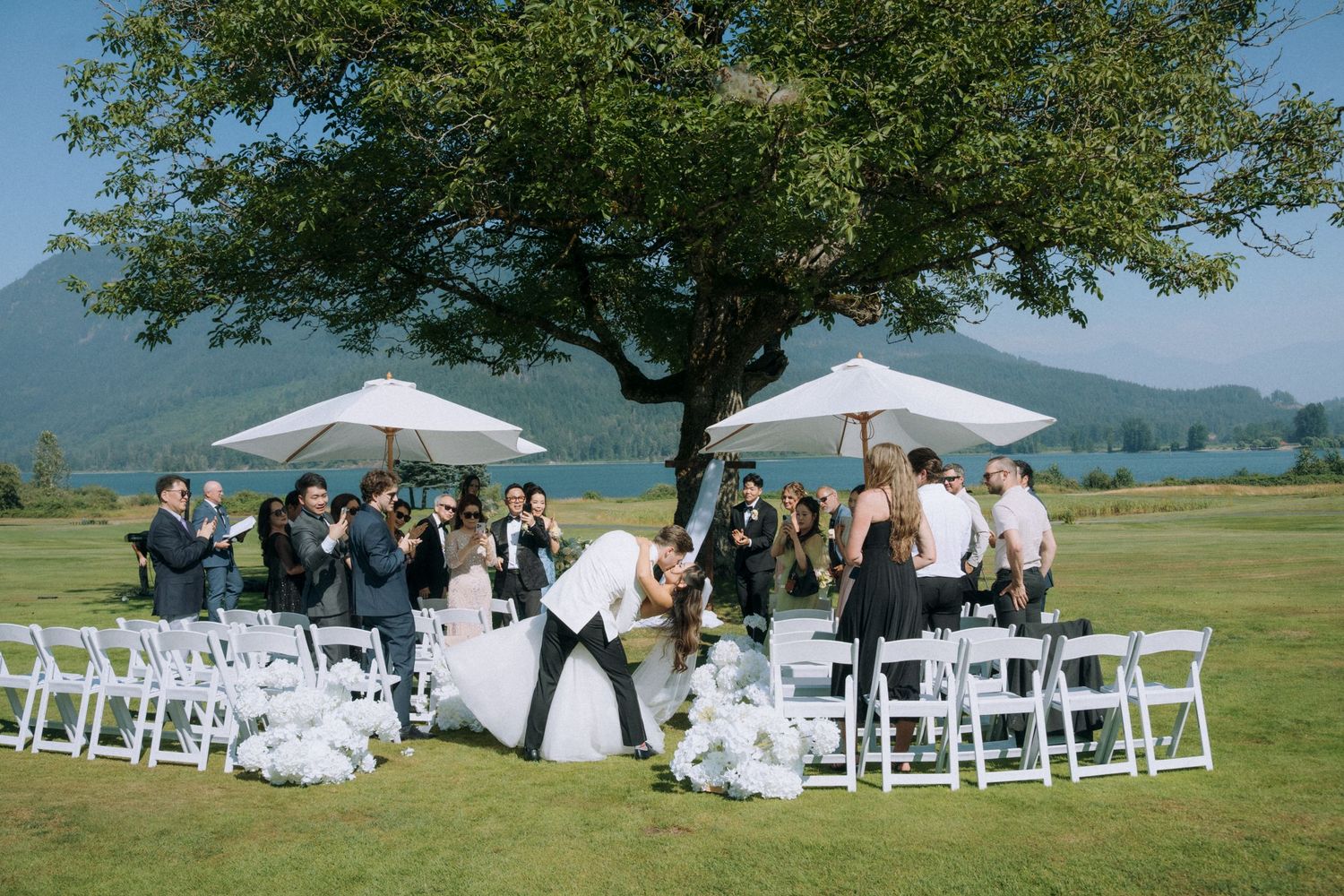 Outdoor wedding ceremony on golf course with couple kissing, white chairs, umbrellas, and mountain lake backdrop.