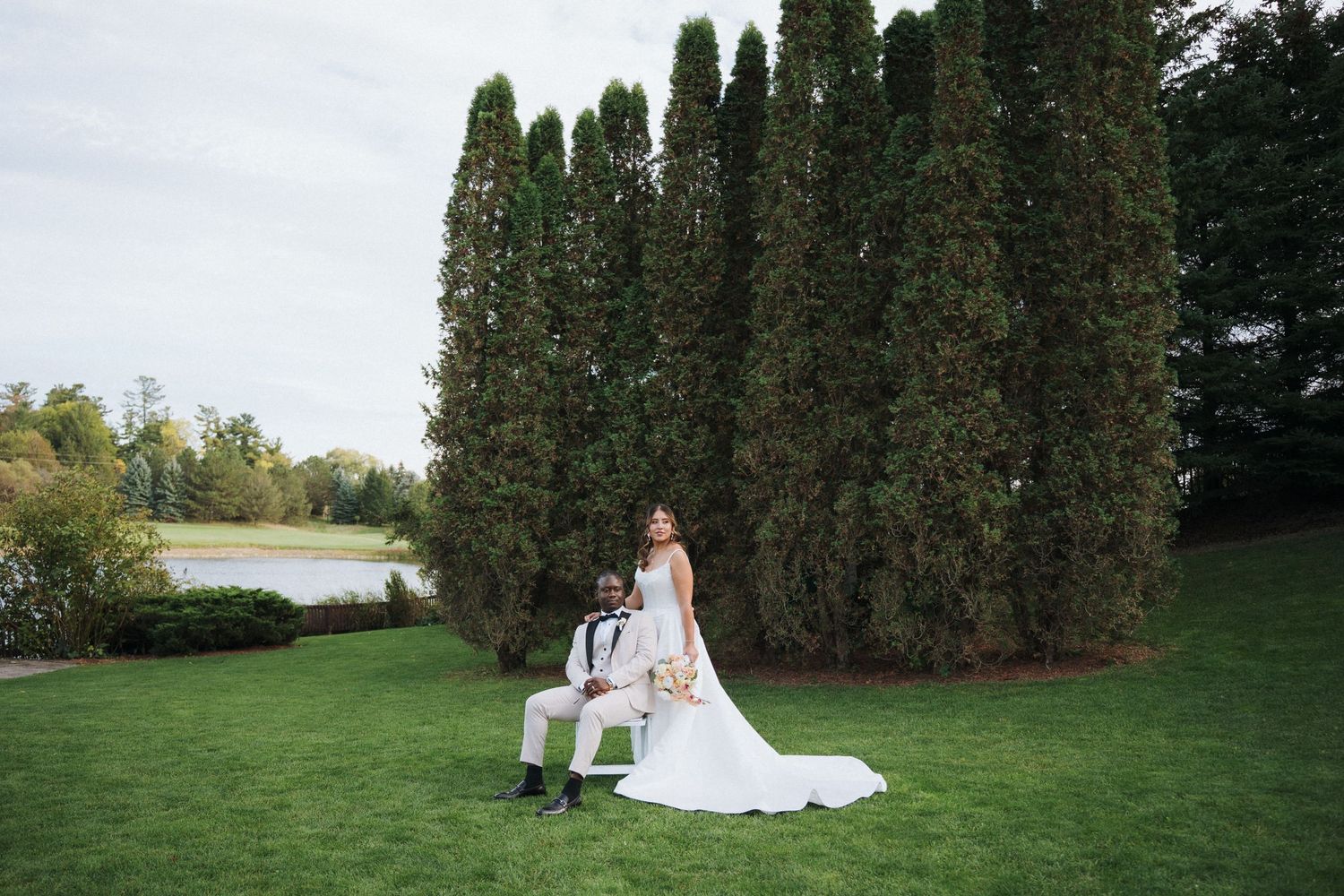 A bride in a white wedding gown poses in a lush garden setting with tall evergreen trees in the background.