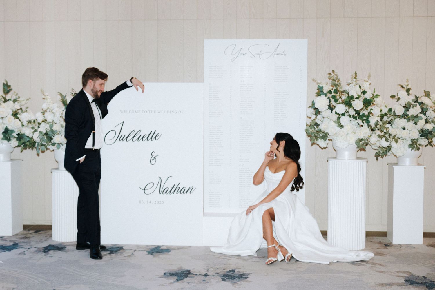 A couple posing playfully with white wedding decorations and floral arrangements against a light colored wall.