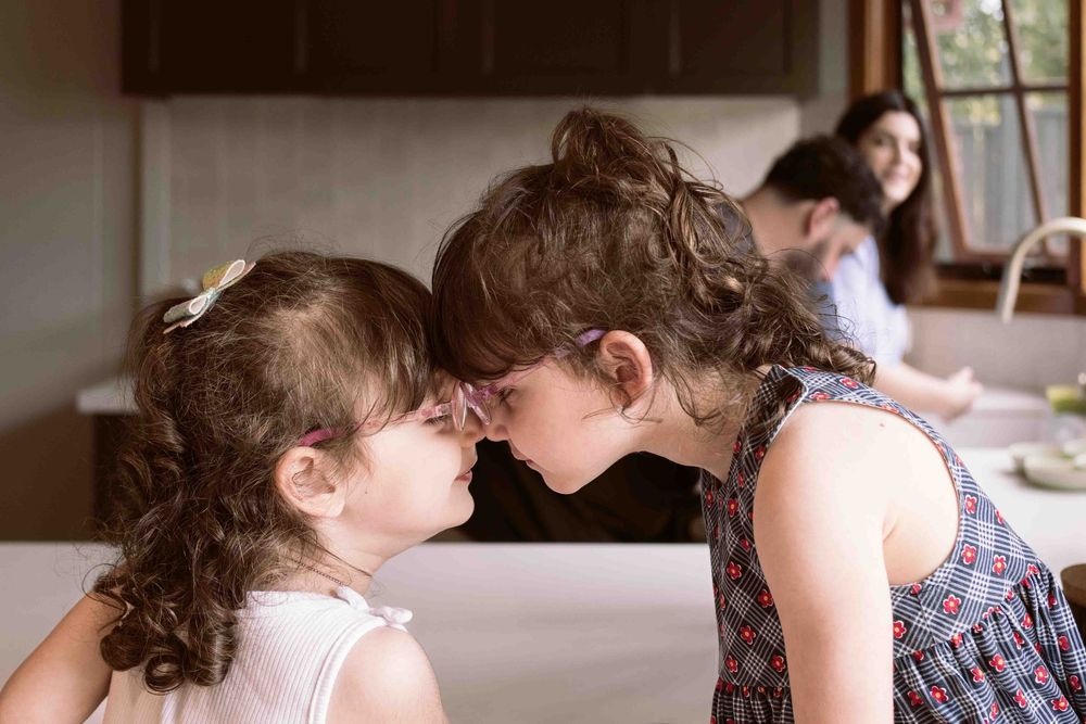 twin girls touching heads together while parents look on from kitchen
