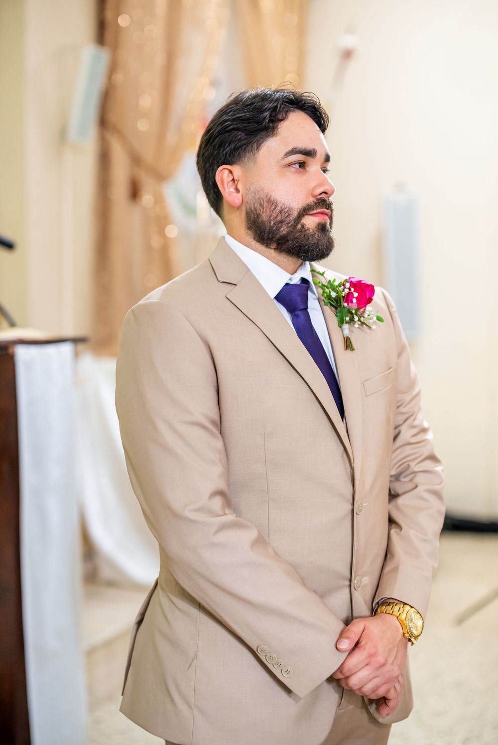 Wedding attendant in a beige suit with navy tie and pink boutonniere at an indoor ceremony venue.