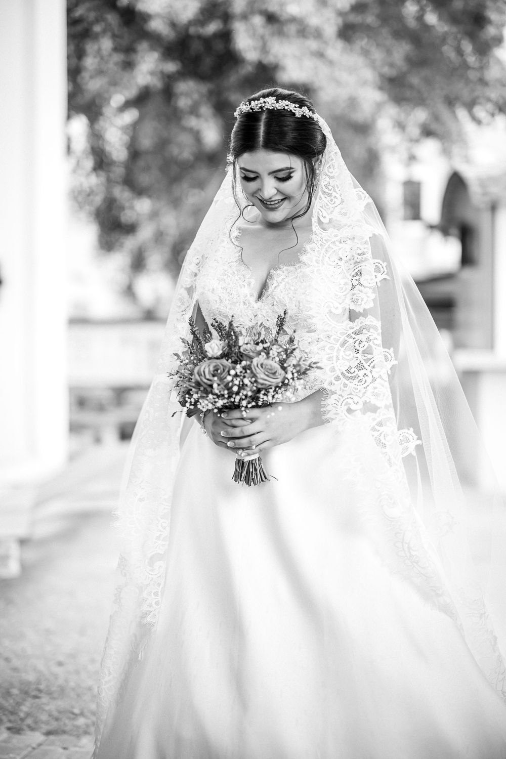 Black and white portrait of a bride in a traditional white lace gown with veil holding a floral bouquet at her wedding.