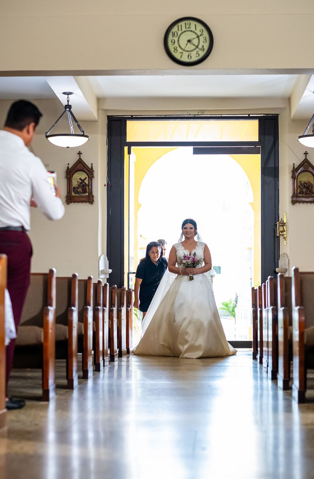 A wedding ceremony processional in a church aisle with bride entering through arched doorway.