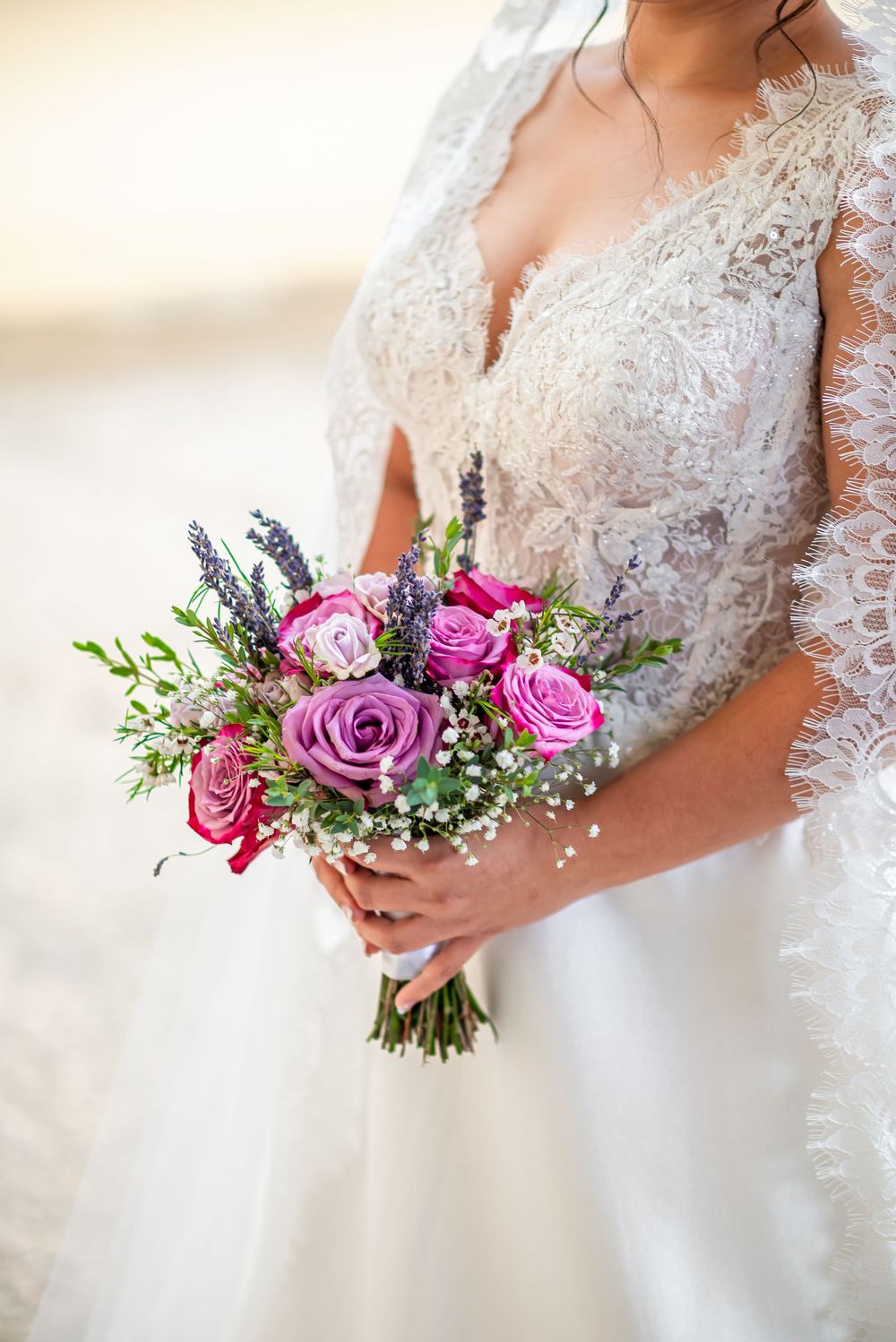 A beautiful bridal bouquet of pink and purple roses with lavender and baby's breath held against a lace wedding dress.