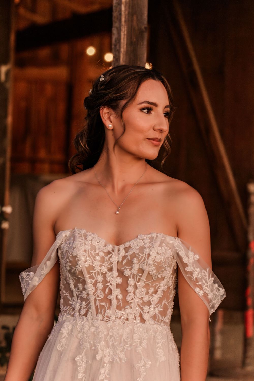 Bride wearing off-shoulder lace wedding gown poses in rustic barn setting.