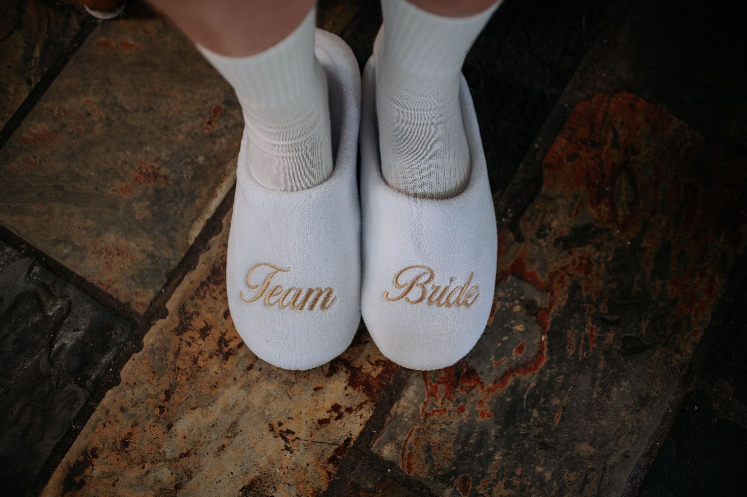 White bridal slippers with 'Team Bride' embroidered in gold text sitting on a rustic stone surface.