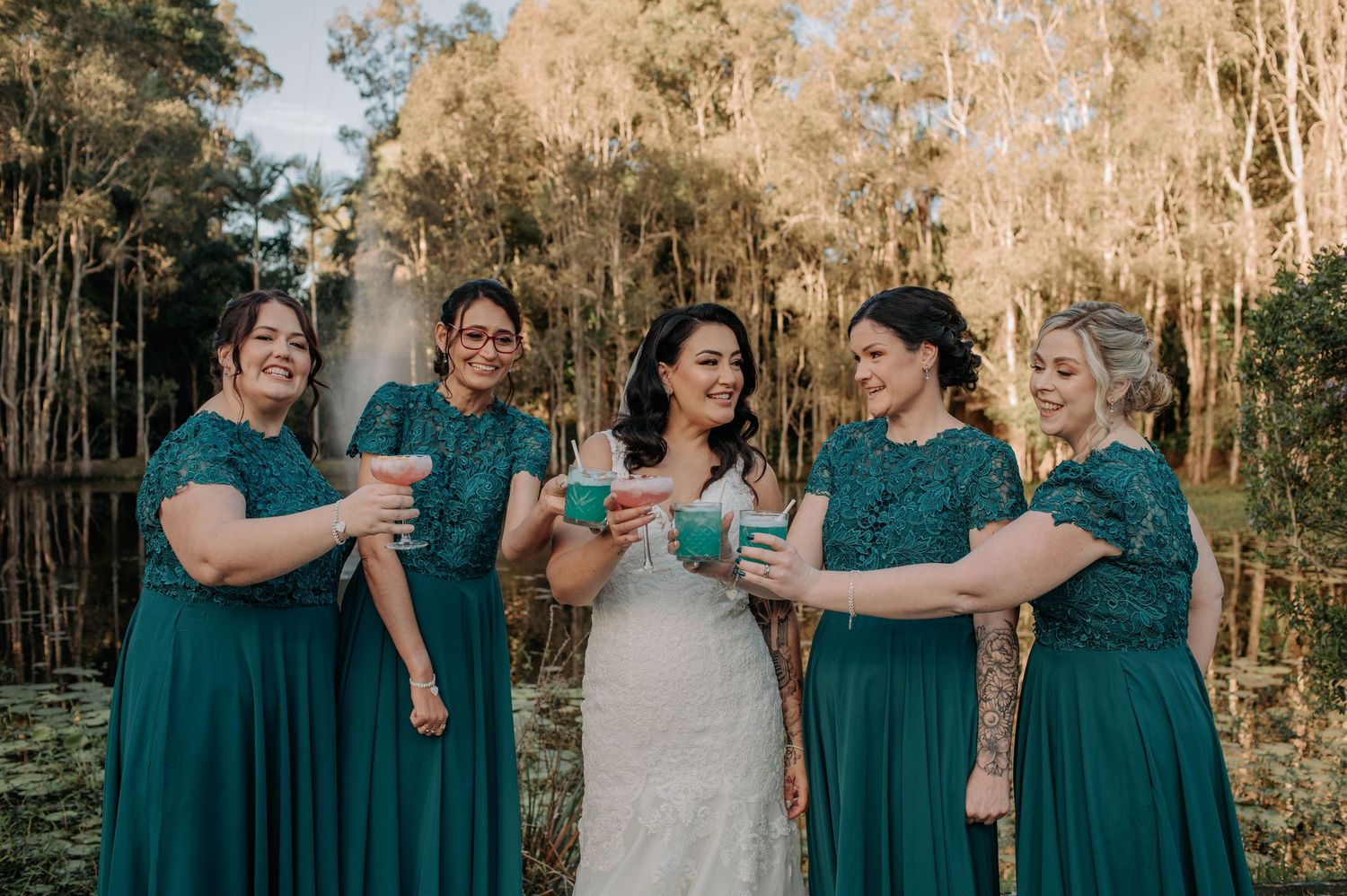 Group of bridesmaids in emerald green dresses sharing champagne toasts with bride in white lace gown at outdoor venue.