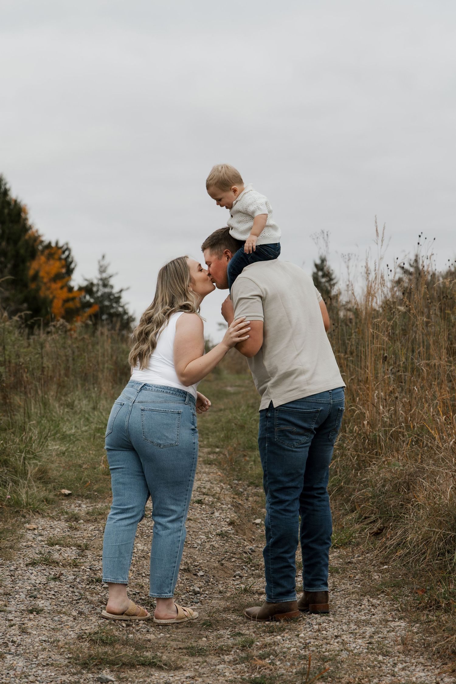 Family portrait on rustic trail with father lifting toddler on shoulders while parents share a kiss outdoors.