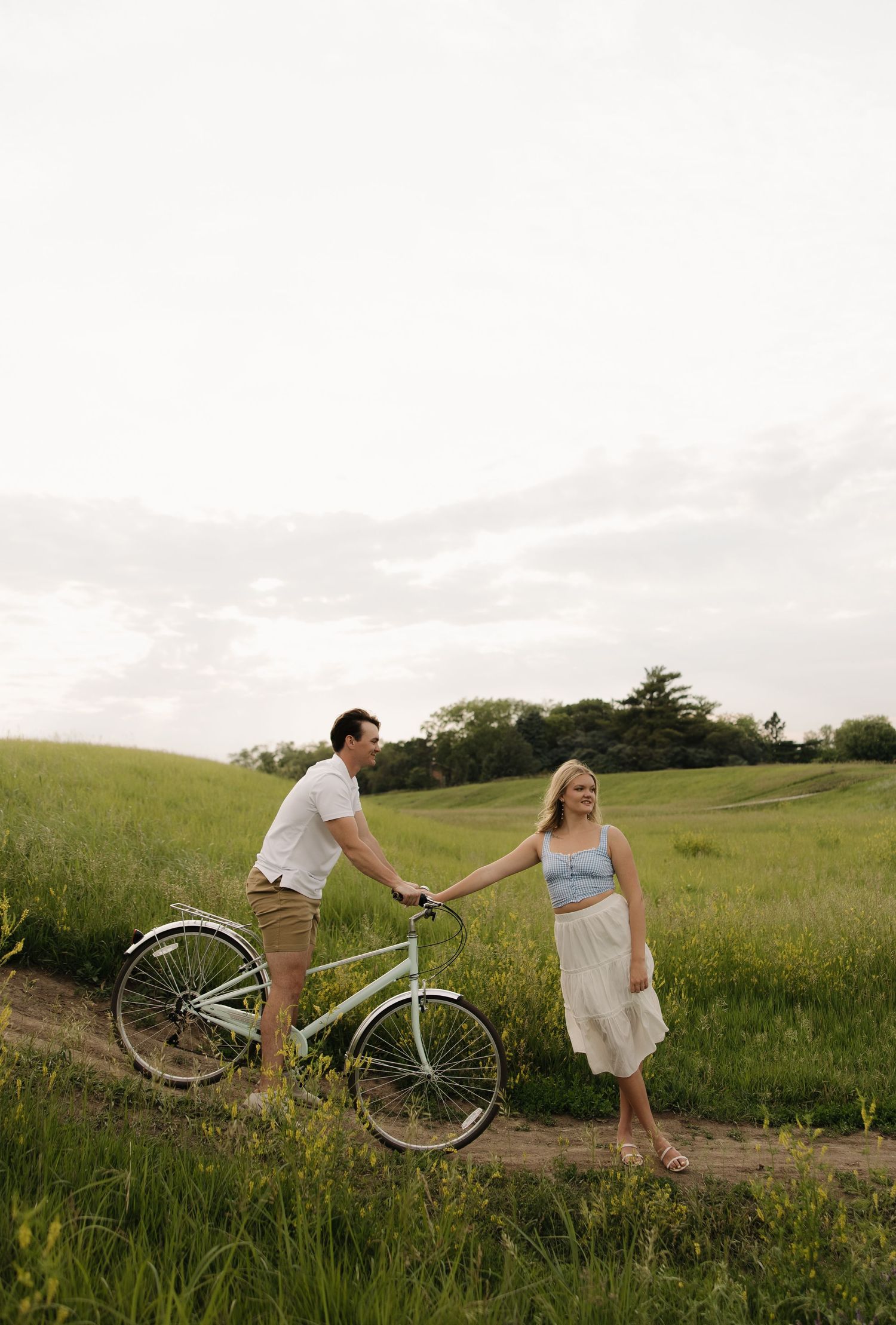 A couple shares a romantic moment on a dirt path in a green meadow at sunset with a vintage bicycle.