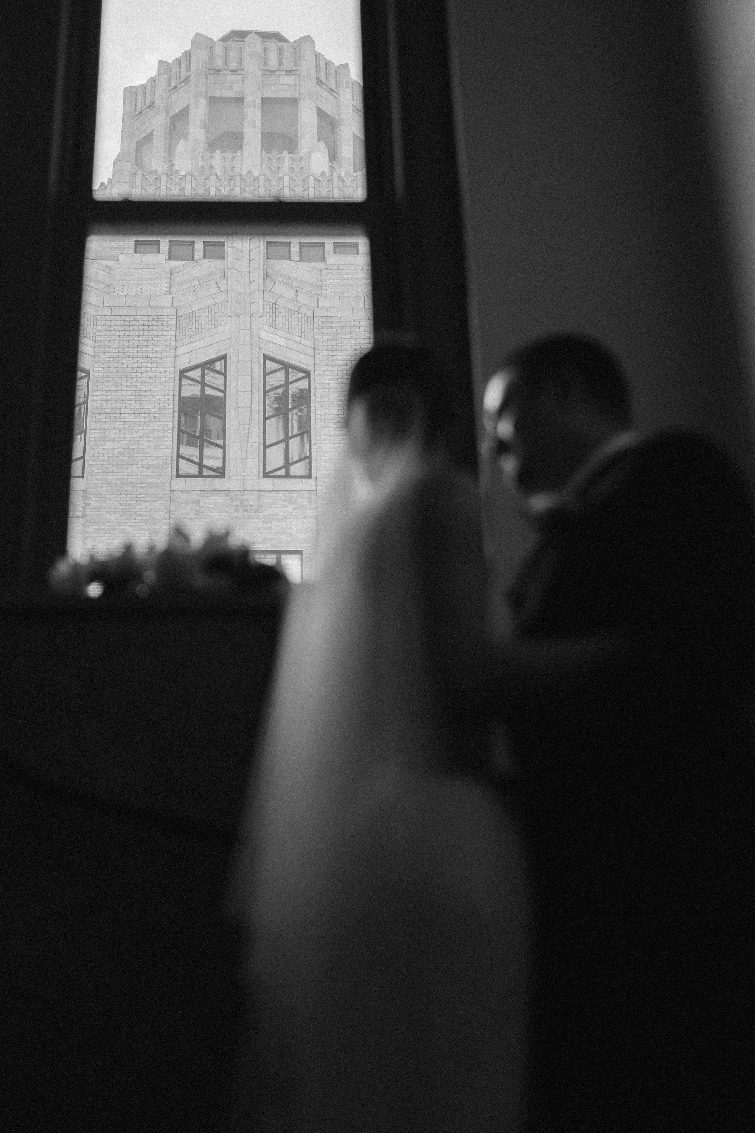 A black and white artistic wedding photograph showing silhouettes against a window with art deco architecture visible outside.